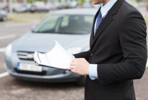 Ein Mann im Anzug hält Vertrag in der Hand. Ein Auto im Hintergrund ist zu sehen.